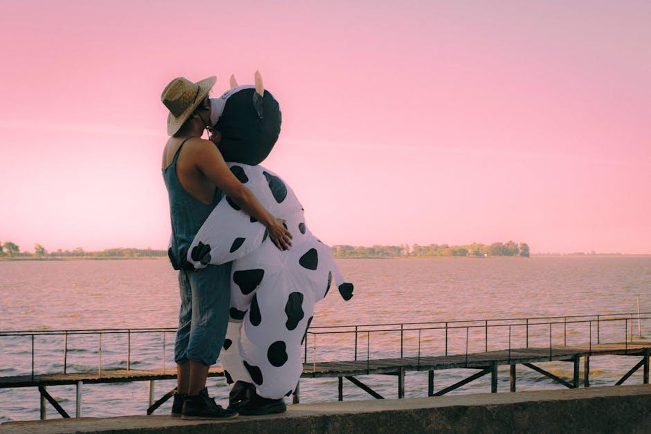 La_Relazione_di_Joe_e_Mika_nel_Tempo_aqYrhKami2 - Confidenze24 Source: Agustina Tolosa - pexels - A couple shares a loving embrace at sunset, with one wearing a fun cow costume by a serene riverside.