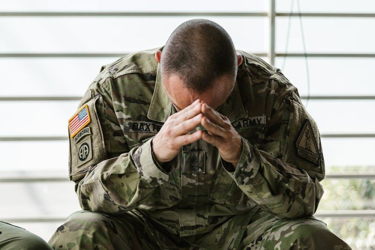 La_controversia_del_bacio__emotivamente_devastante_per_la_moglie_Georgia_rG3JVJYAy8 - Confidenze24 Source: RDNE Stock project - pexels - A soldier in camouflage sitting indoors, appearing stressed and overwhelmed.