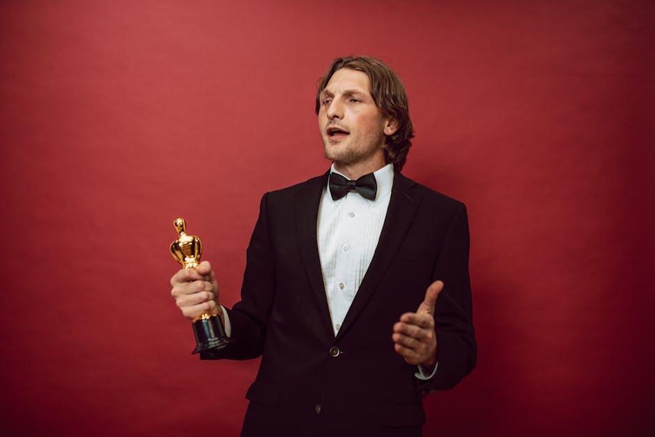 La_dedica_a_marito_e_figli_nel_discorso_di_accettazione_NCGXmzfmyK - Confidenze24 Source: RDNE Stock project - pexels - An accomplished man in a tuxedo holding a trophy, symbolizing triumph and recognition, against a red backdrop.
