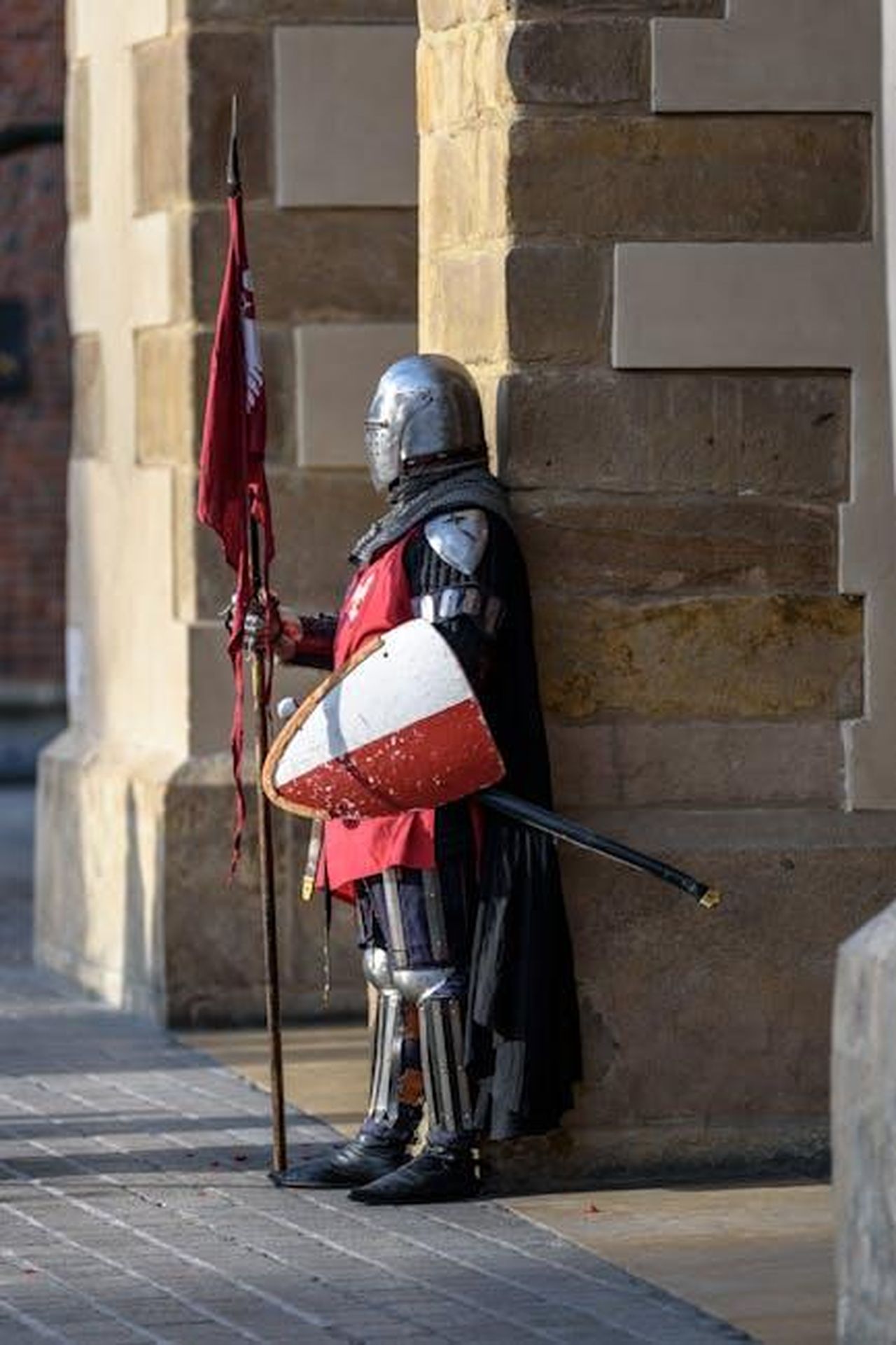 La_difesa_di_Errol_Musk_txgaTtZpD4 - Confidenze24 Source: AS Photography - pexels - A medieval knight in armor stands guard with a shield and flag outside a stone building.