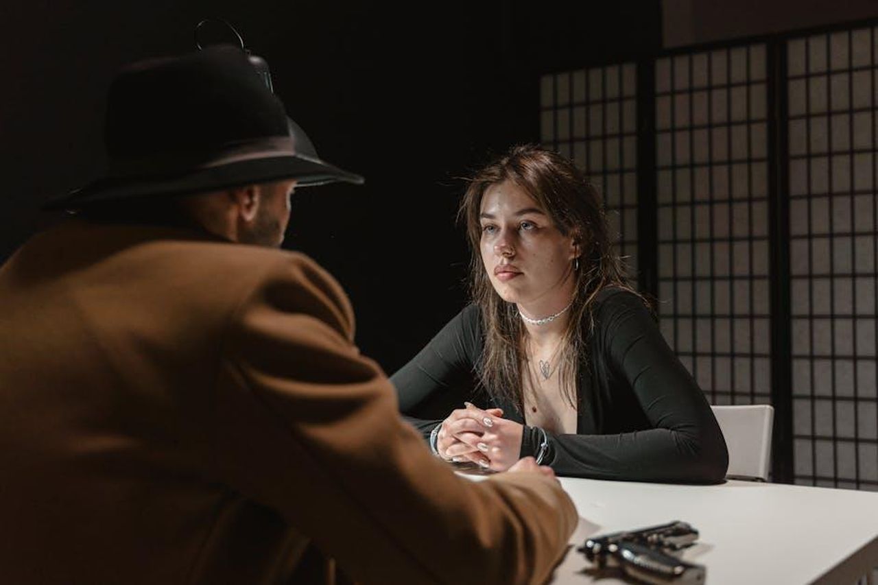 La_fine_di_unera_14_stagioni_di_Blue_Bloods_EQ8YS9Caoj - Confidenze24 Source: MART PRODUCTION - pexels - A woman faces a man in a hat during a tense interrogation scene at a table.