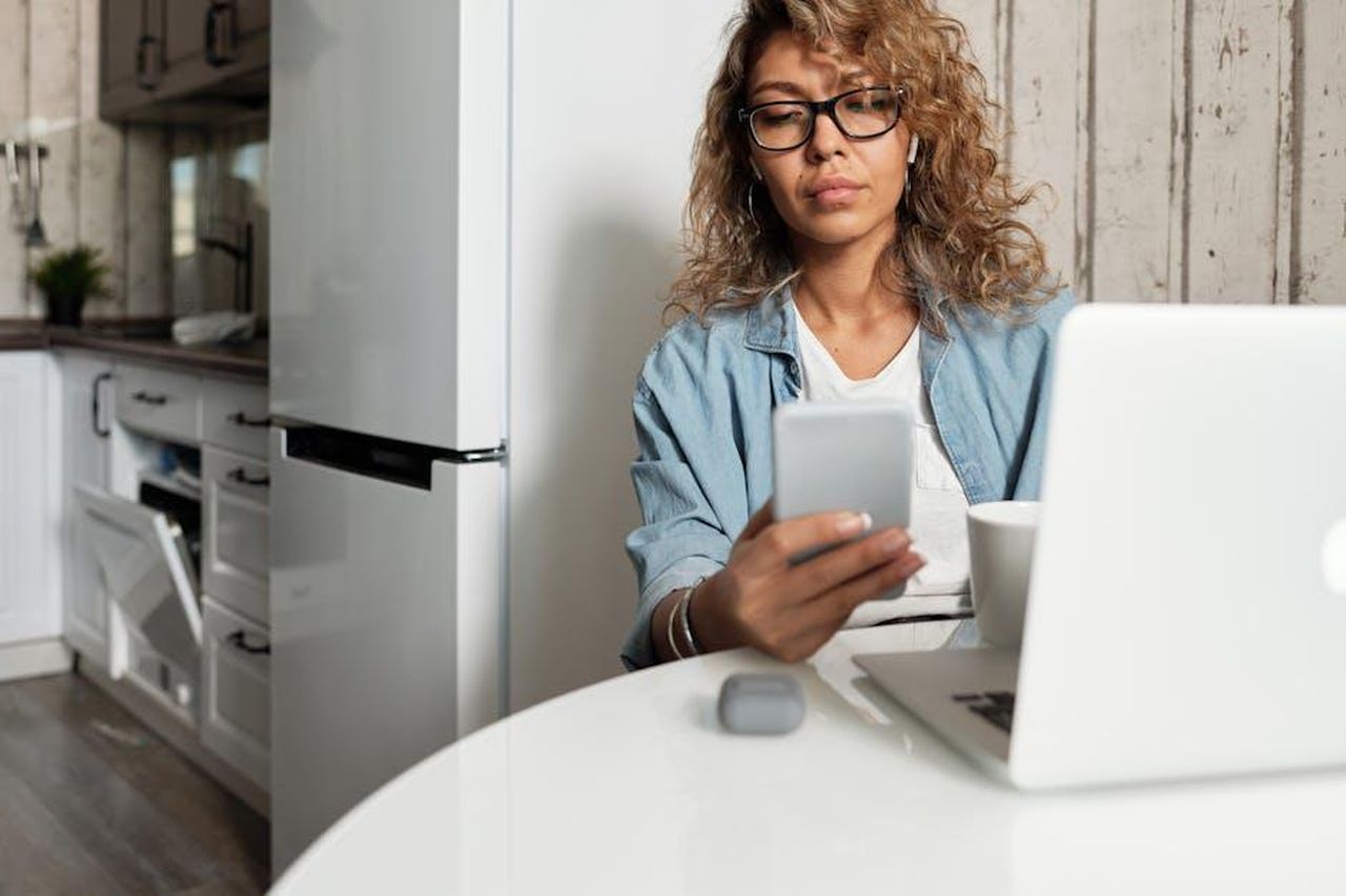 La_newsletter_di_As_Ever_kGpi8F5wXn - Confidenze24 Source: Matilda Wormwood - pexels - Woman using smartphone and laptop while working remotely in a home kitchen setting.
