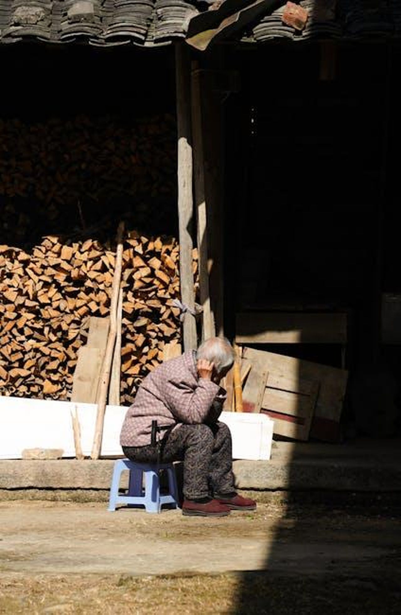 La_pausa_di_Nicole_Kidman_1gfC2nUFGA - Confidenze24 Source: Ban Huang - pexels - An elderly person sits on a stool near a stack of firewood on a sunny day outdoors.