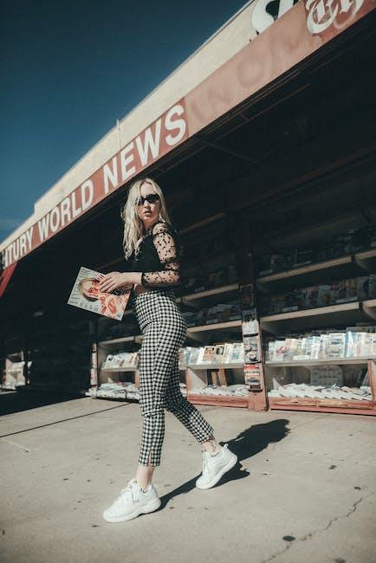 La_reazione_di_Jennifer_Lopez_e_le_voci_su_Ben_Affleck_OXmh9j8VvR - Confidenze24 Source: Nina Hill - pexels - Stylish woman in black lace top walking past newsstand holding a magazine, under bright sunlight.
