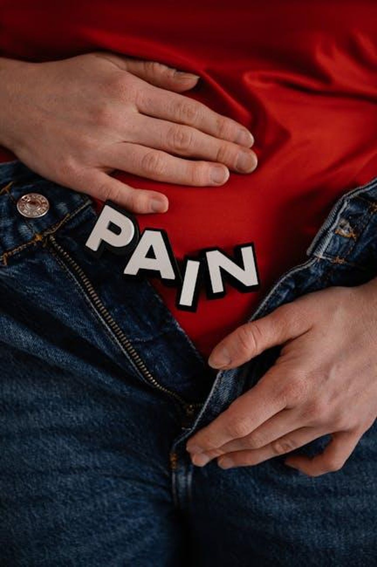 La_risposta_del_produttore_Jesse_Fawcett_RMeQAWchNw - Confidenze24 Source: SHVETS production - pexels - Close-up of a person in jeans holding their stomach with 'PAIN' text overlay.