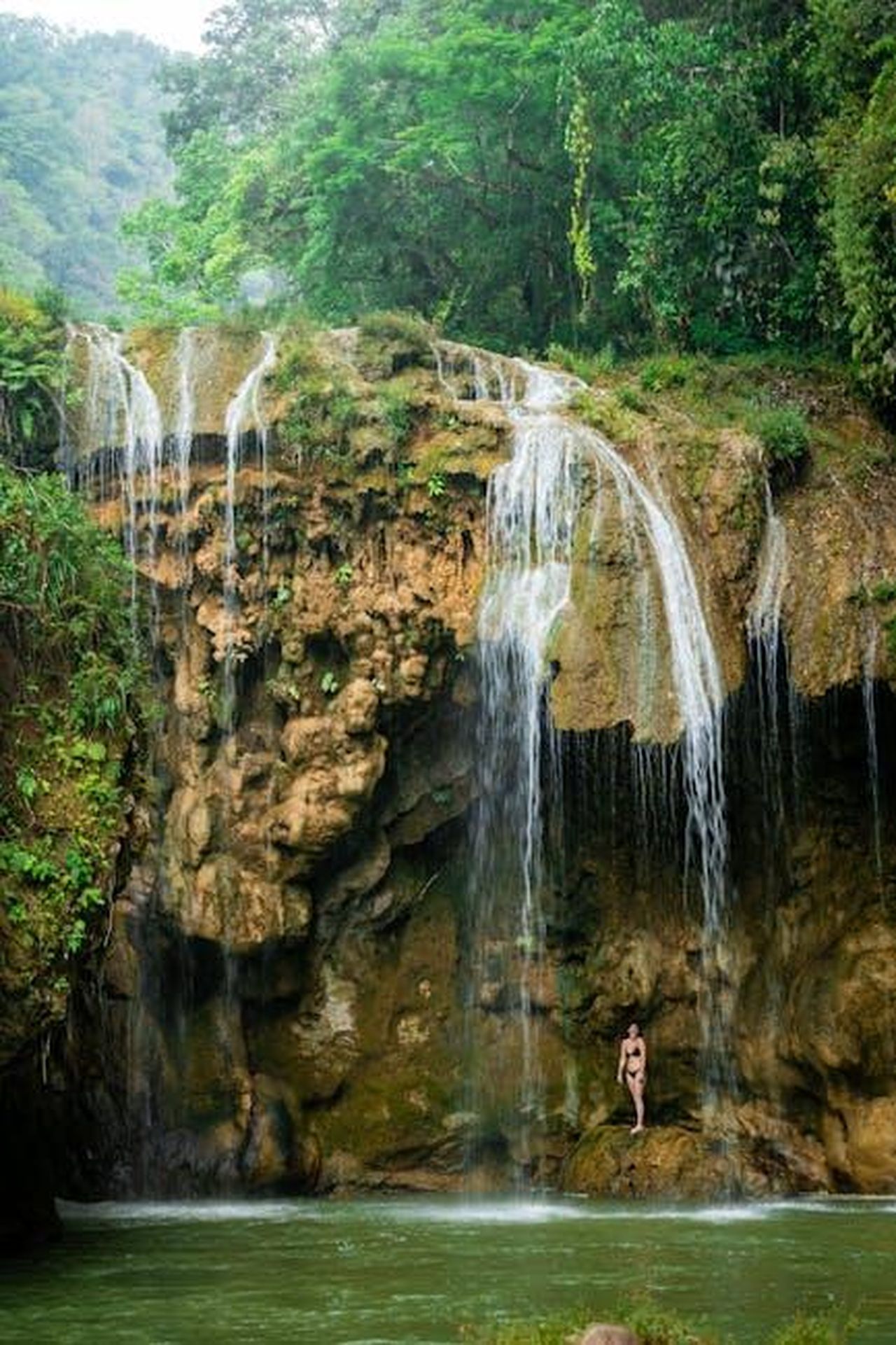 La_risposta_di_Zoe_Saldaa_alle_critiche_qKNph67nqT - Confidenze24 Source: Diego Girón - pexels - A woman stands near a stunning waterfall in Mae Ping National Park, surrounded by lush greenery.