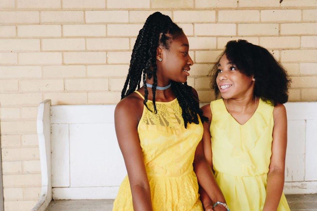 La_scoperta_dei_suoi_fratelli_e_sorelle_oSFwRWc47w - Confidenze24 Source: nappy - pexels - Two sisters in yellow dresses smiling at each other, radiating joy indoors.