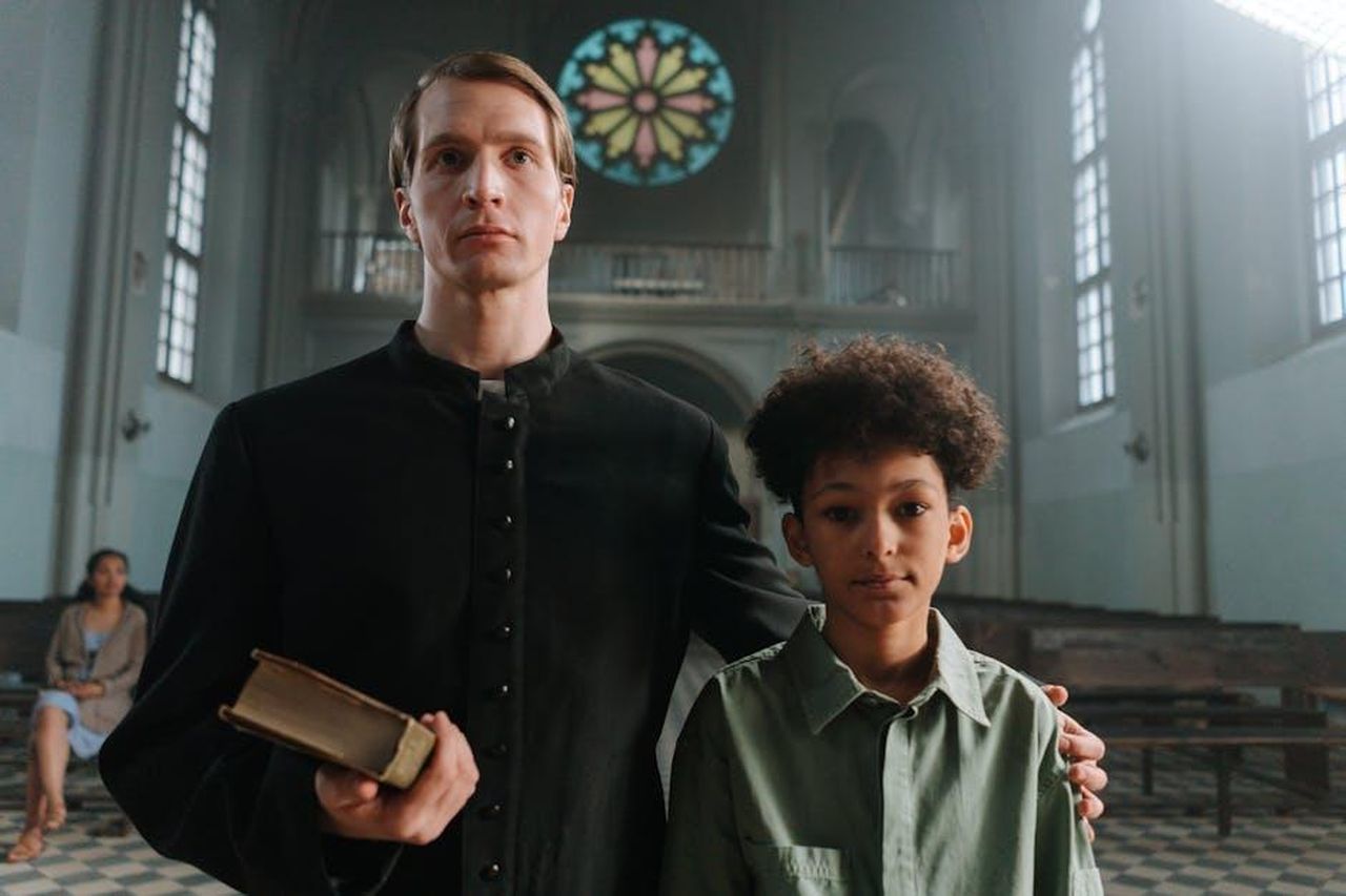 La_storia_delle_relazioni_di_Gethin_SXL6RyCfjc - Confidenze24 Source: cottonbro studio - pexels - A priest standing with a young boy in a church featuring a colorful stained glass window.
