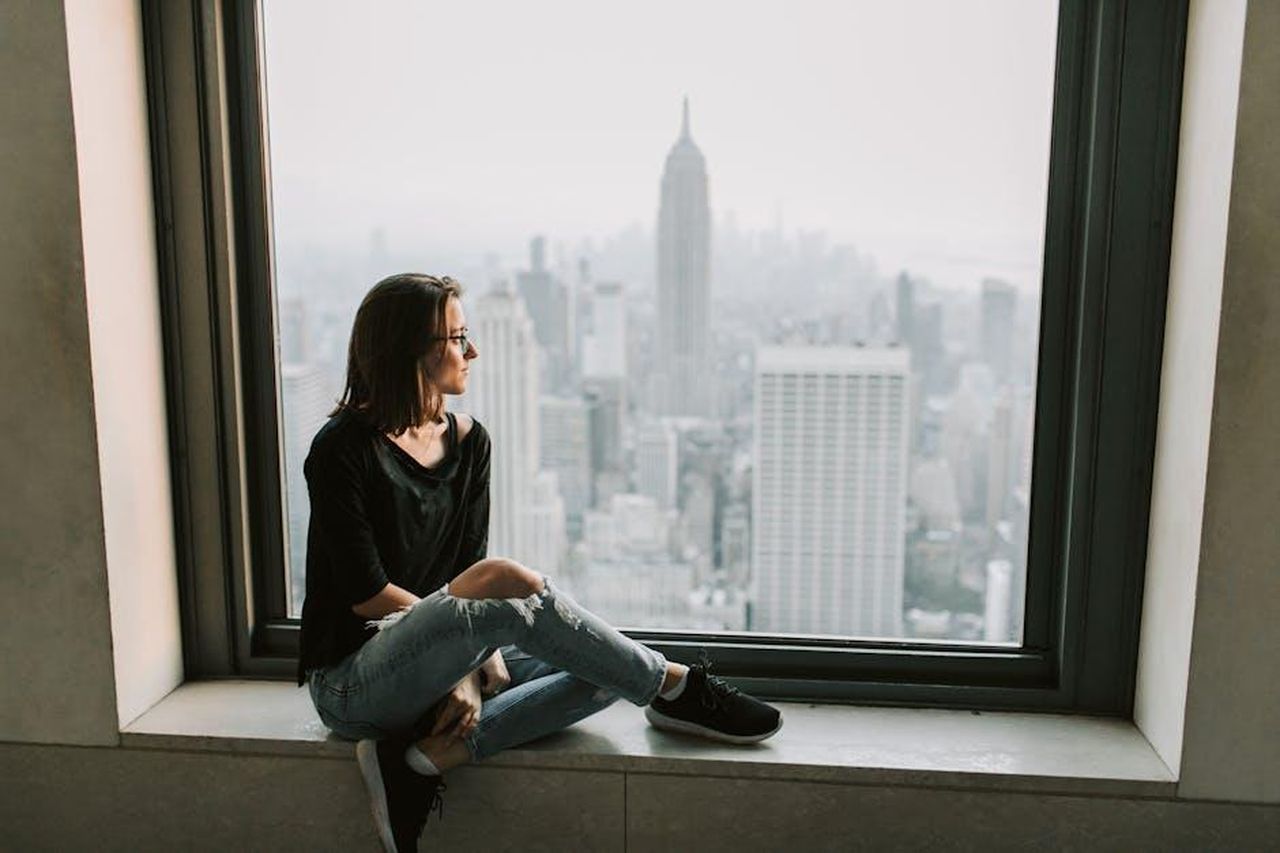 La_vita_di_George_e_Amal_a_New_York_Prxj3bUmlC - Confidenze24 Source: Vlada Karpovich - pexels - A woman sits on a windowsill, admiring the New York City skyline with the Empire State Building in view.