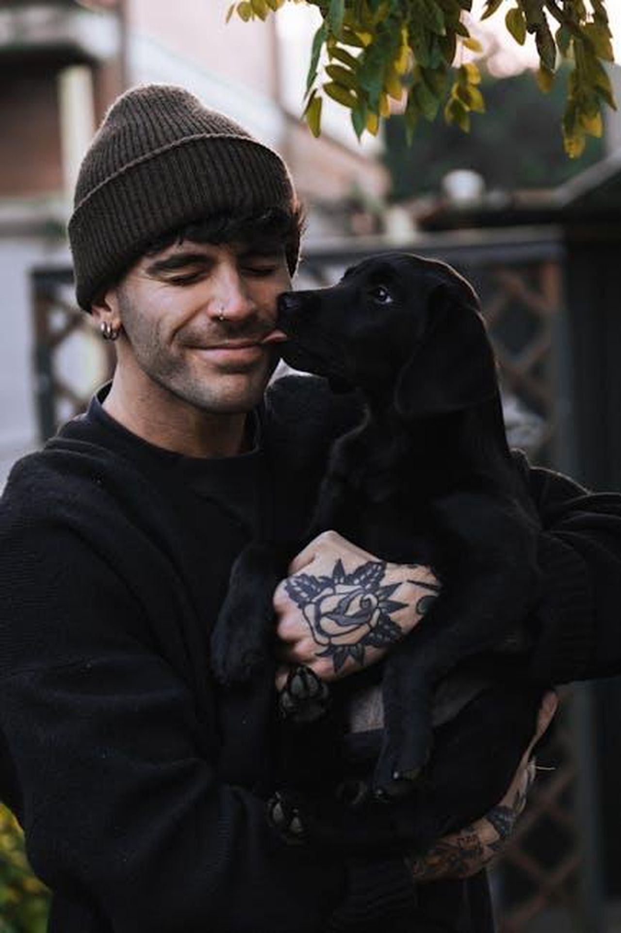 La_vita_di_Pierce_Brosnan_e_la_sua_famiglia_t99DiyFcyE - Confidenze24 Source: Stefania Spadoni - pexels - Smiling man with tattoos holding a playful black Labrador puppy outdoors.