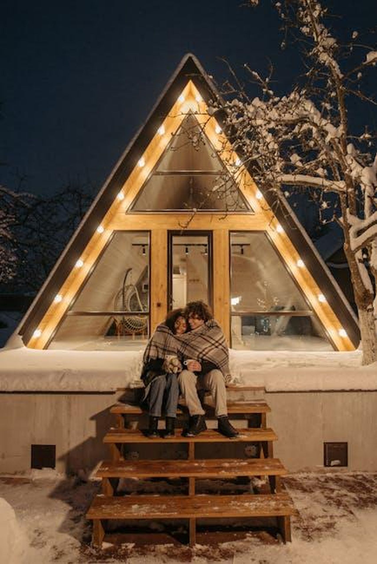 La_vita_insieme_di_Amy_e_TJ_abitudini_e_sorprese_JmDu48bHQx - Confidenze24 Source: Pavel Danilyuk - pexels - A couple sits warmly wrapped on steps of an A-frame cabin on a snowy winter night, enjoying a romantic moment.
