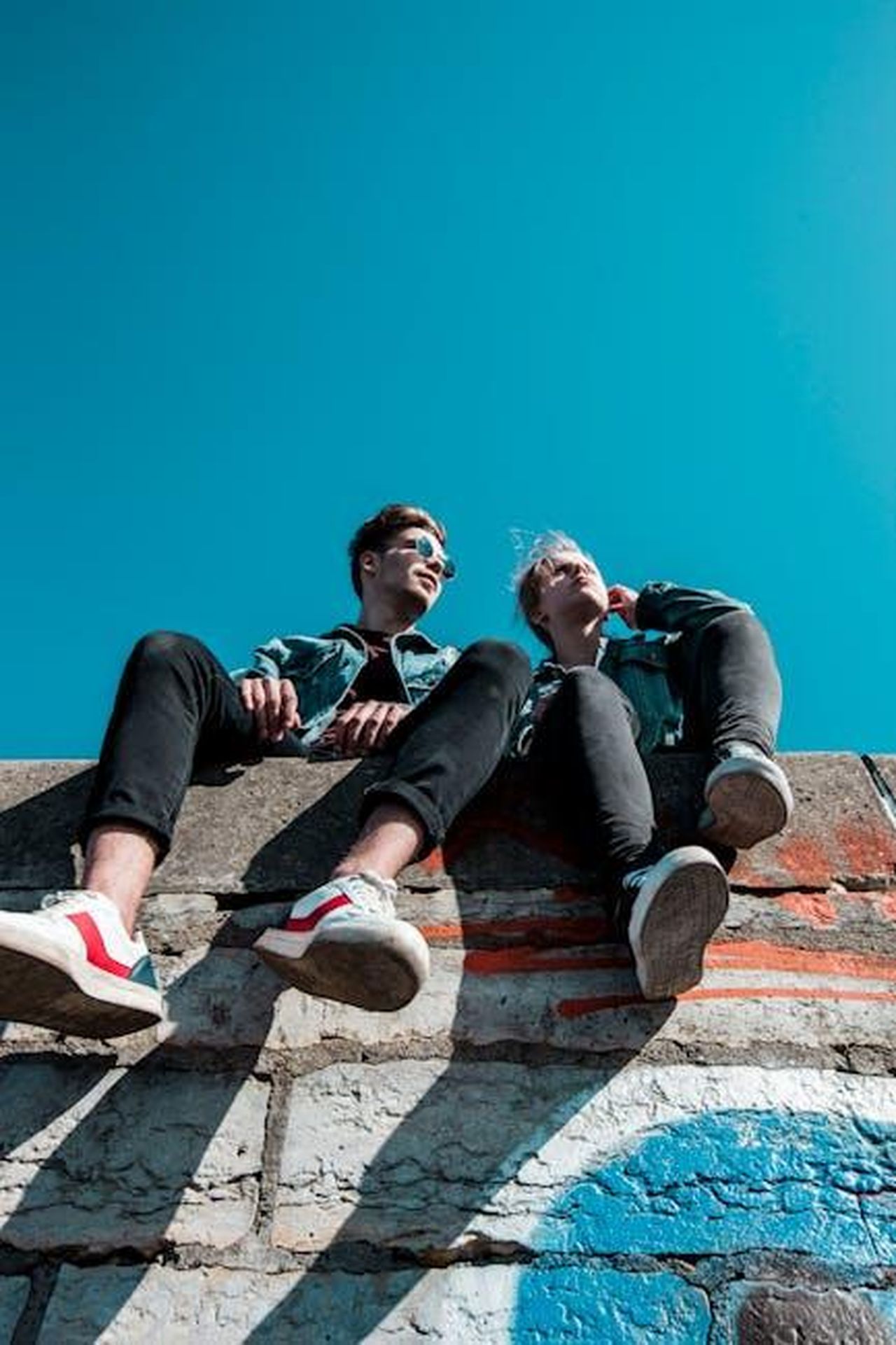 Lamicizia_tra_Danny_e_Tom__in_pericolo_jJwl3FYR7m - Confidenze24 Source: Marlene Leppänen - pexels - Two young people sitting on a stone wall under a vibrant blue sky, enjoying a sunny day together.