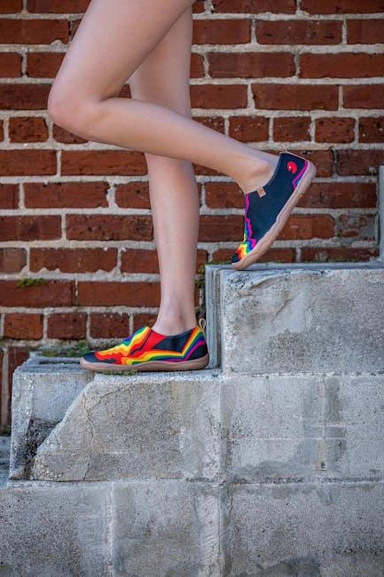 Lancio_della_collezione_Sperry_x_Aritzia_xw3v2GcfVN - Confidenze24 Source: Connor McManus - pexels - Vibrant rainbow sneakers worn by woman on concrete steps against brick wall.