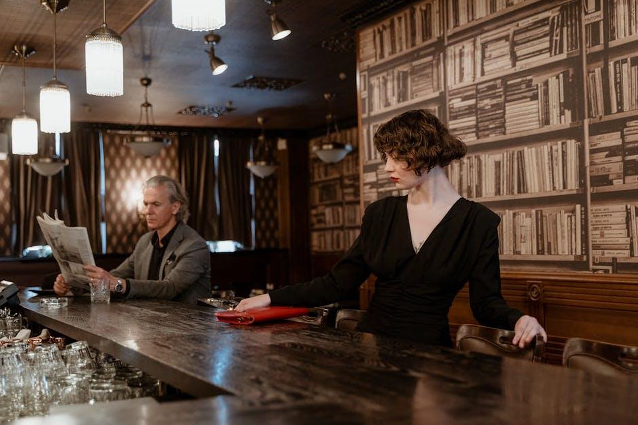 Le_Dichiarazioni_di_Tamar_Braxton_sul_Ristorante_Old_Lady_Gang_STRlSdttNR - Confidenze24 Source: MART PRODUCTION - pexels - Stylish indoor scene with a man reading a newspaper and a woman in a black dress at a bar.
