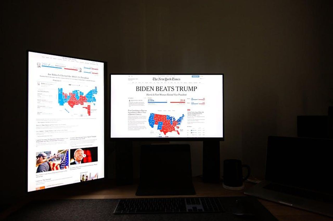 Le_accuse_contro_Harry_e_la_reazione_dei_media_8jj9PaT4s4 - Confidenze24 Source: Andrew Neel - pexels - Dual computer screens in a dark room display election results indicating Biden's victory over Trump.