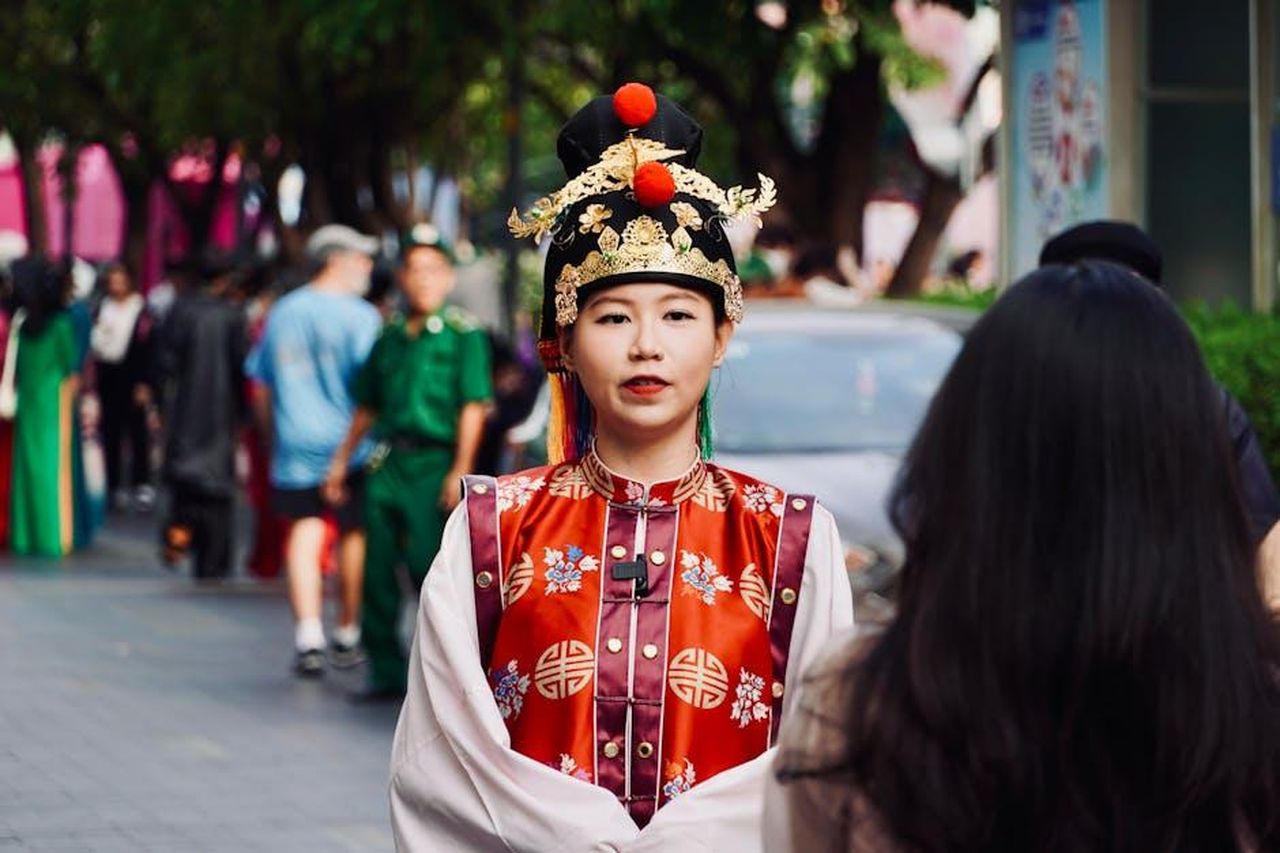 Le_conseguenze_della_sua_estromissione_Tdp0hzv6CM - Confidenze24 Source: Tuan Vy - pexels - Woman in ornate traditional Asian dress and headdress captured at an outdoor event with blurred people in background.