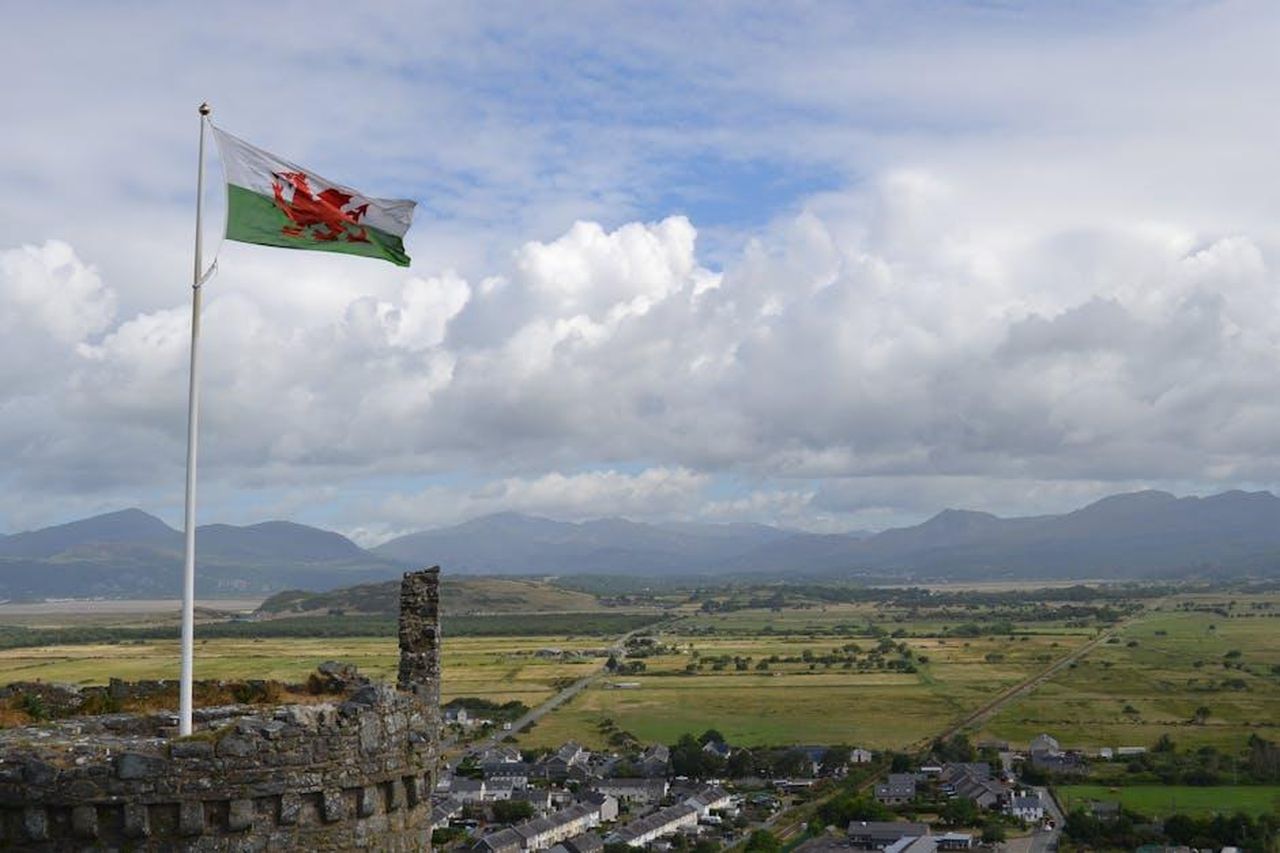 Le_critiche_del_professor_Evans_ZXtCvUjfHk - Confidenze24 Source: Leo Wharfe - pexels - A Welsh flag waves over a scenic countryside landscape with mountains and a town visible.