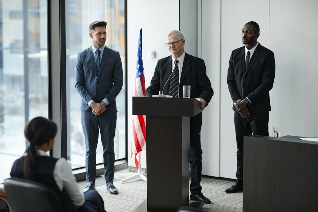 Le_dichiarazioni_degli_ufficiali_americani_FBKgjYq4xw - Confidenze24 Source: Werner Pfennig - pexels - Officials delivering a political speech in a modern conference room with an American flag.