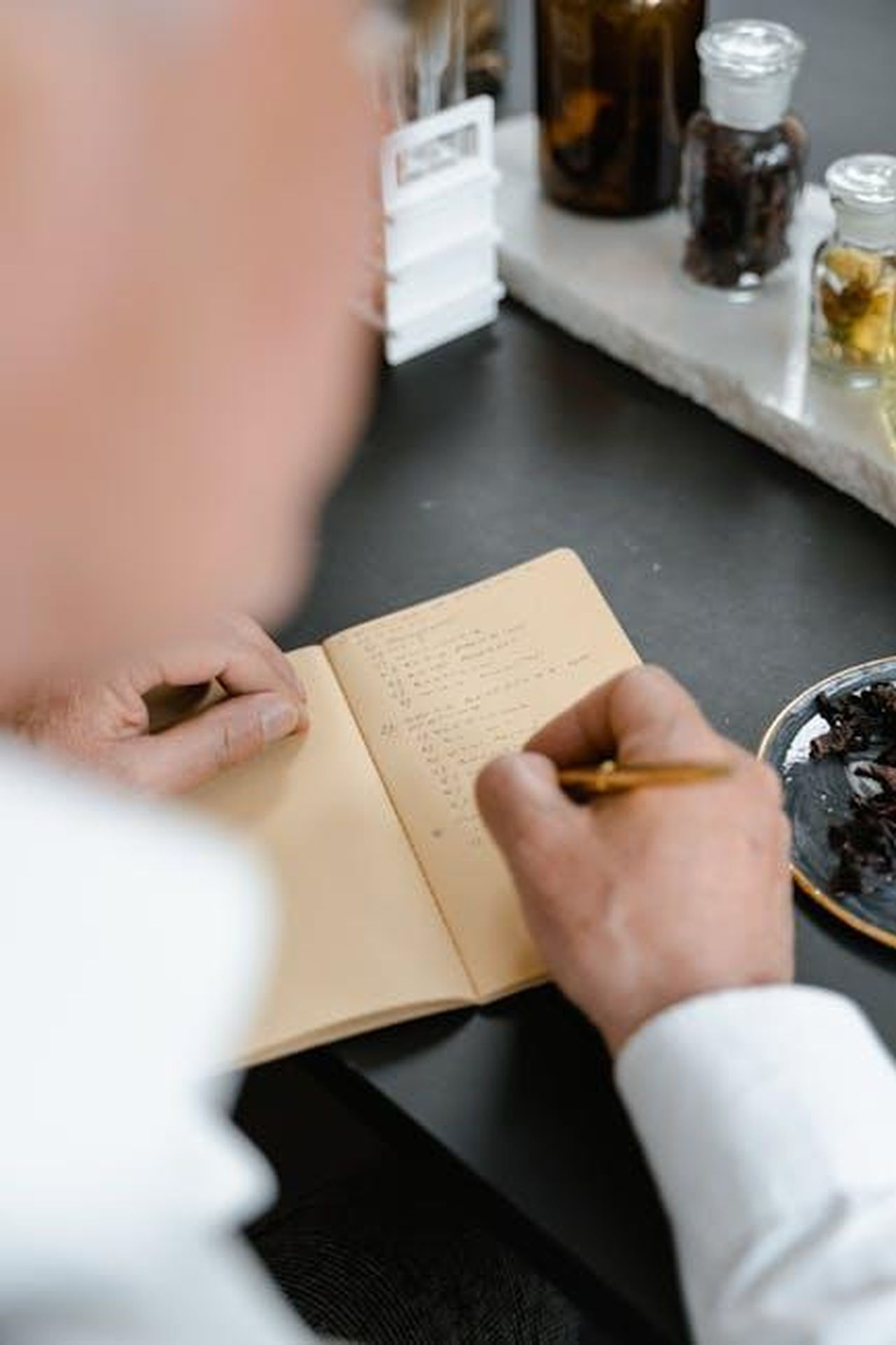 Le_note_olfattive_di_Guerlain_Shalimar_nMWenpmJOh - Confidenze24 Source: MART PRODUCTION - pexels - A person writes notes in a notebook next to aromatherapy ingredients.