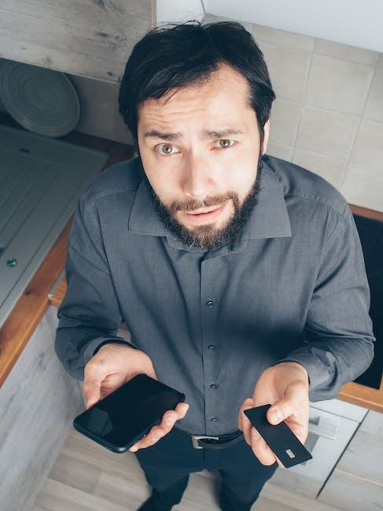 Le_preoccupazioni_per_la_sicurezza_di_Camilla_xSztqnd3RR - Confidenze24 Source: Mikhail Nilov - pexels - A confused man indoors looks up while holding a smartphone and credit card, concerned about a financial issue.