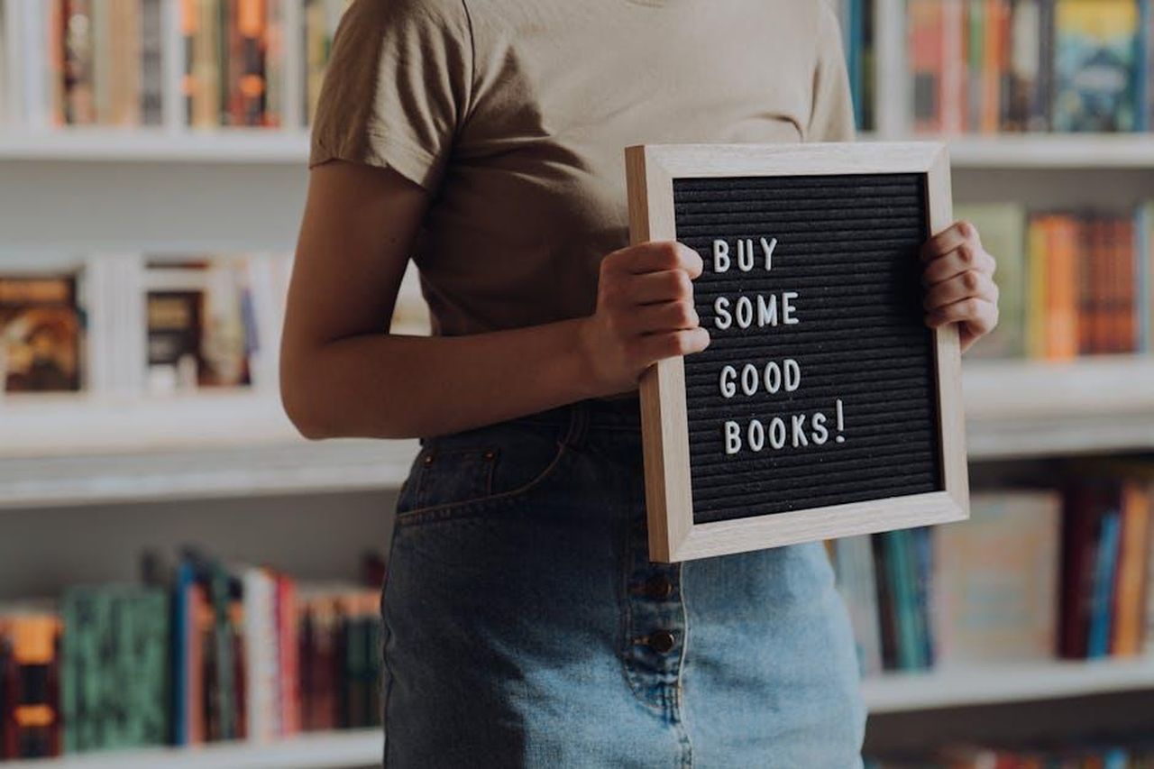 Le_raccomandazioni_di_Reese_Witherspoon_e_Jenna_Bush_Hager_UQId3EvW0V - Confidenze24 Source: cottonbro studio - pexels - A person holding a board with 'Buy Some Good Books' in a bookstore setting.