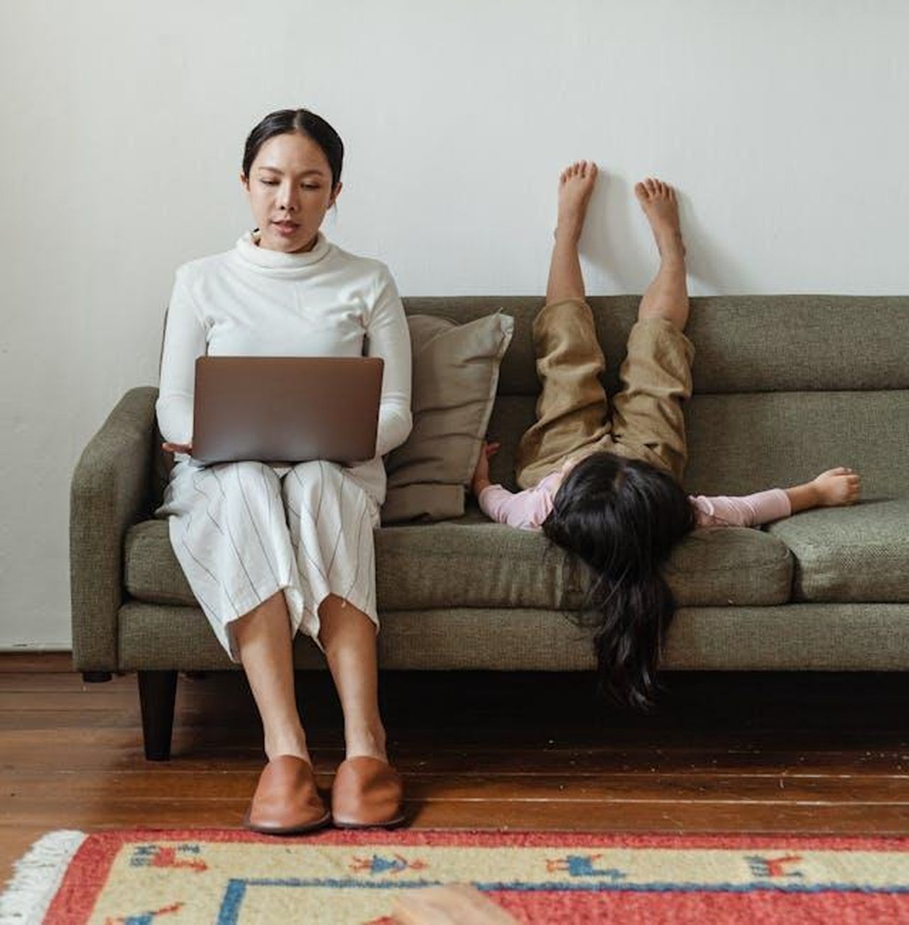 Le_sfide_della_maternit_durante_il_divorzio_Q9v2QjndUn - Confidenze24 Source: Ketut Subiyanto - pexels - Mother multitasking with laptop while daughter plays on sofa, balancing work and family.