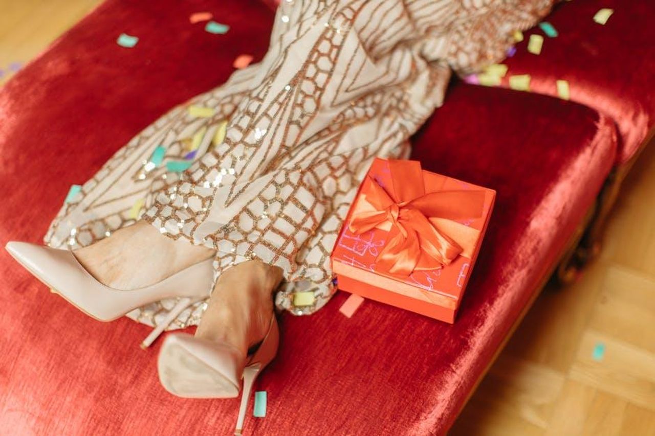 Le_ultime_notizie_su_celebrit_e_cultura_pop_BUgl4WuJzE - Confidenze24 Source: Anastasia Shuraeva - pexels - A stylish scene with high heels, a wrapped gift, and colorful confetti on a red sofa.