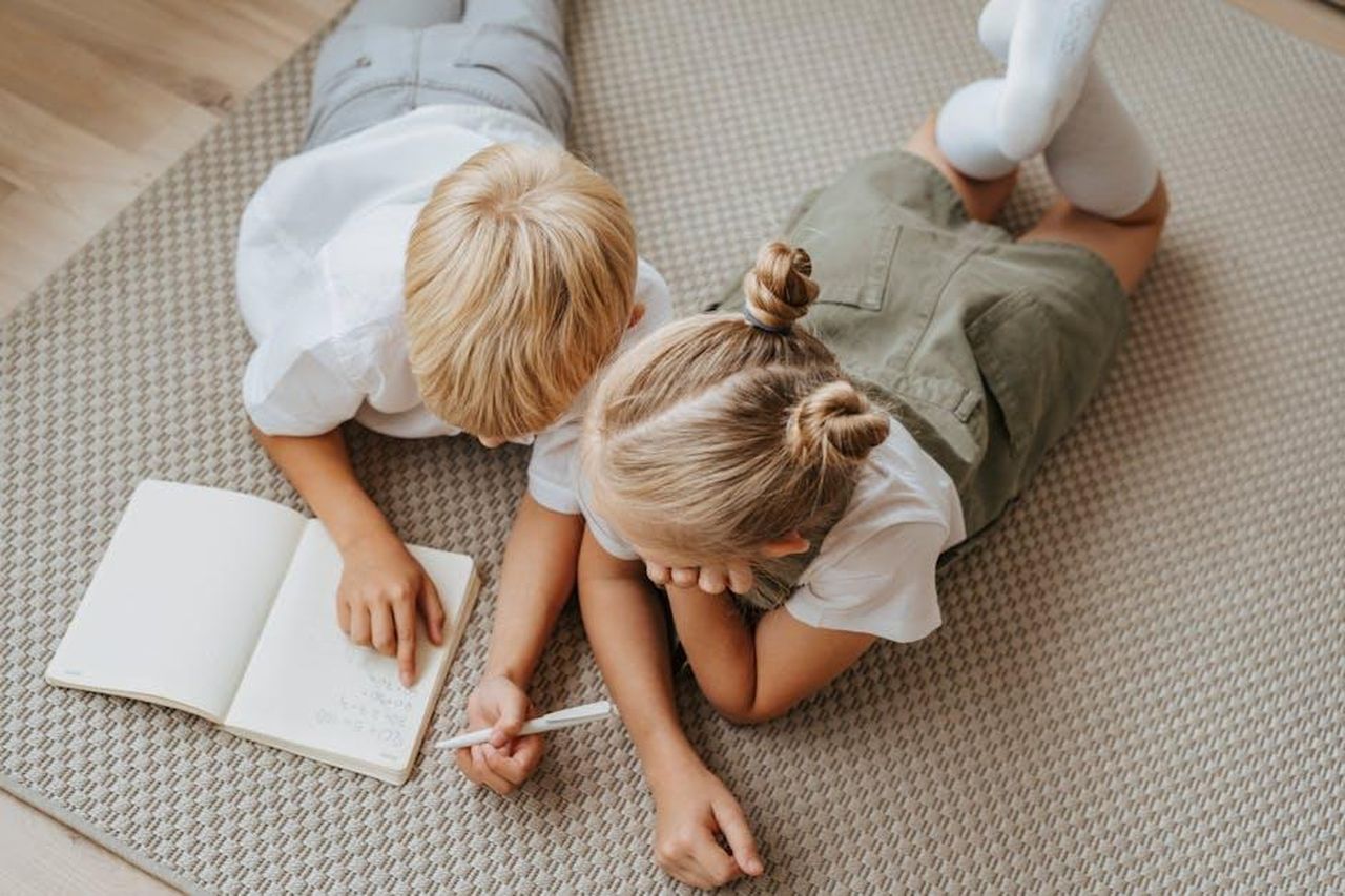 Legame_tra_fratelli_4LxyuGbfaH - Confidenze24 Source: olia danilevich - pexels - Two young children study together on the floor, concentrating on a notebook in a warm indoor setting.