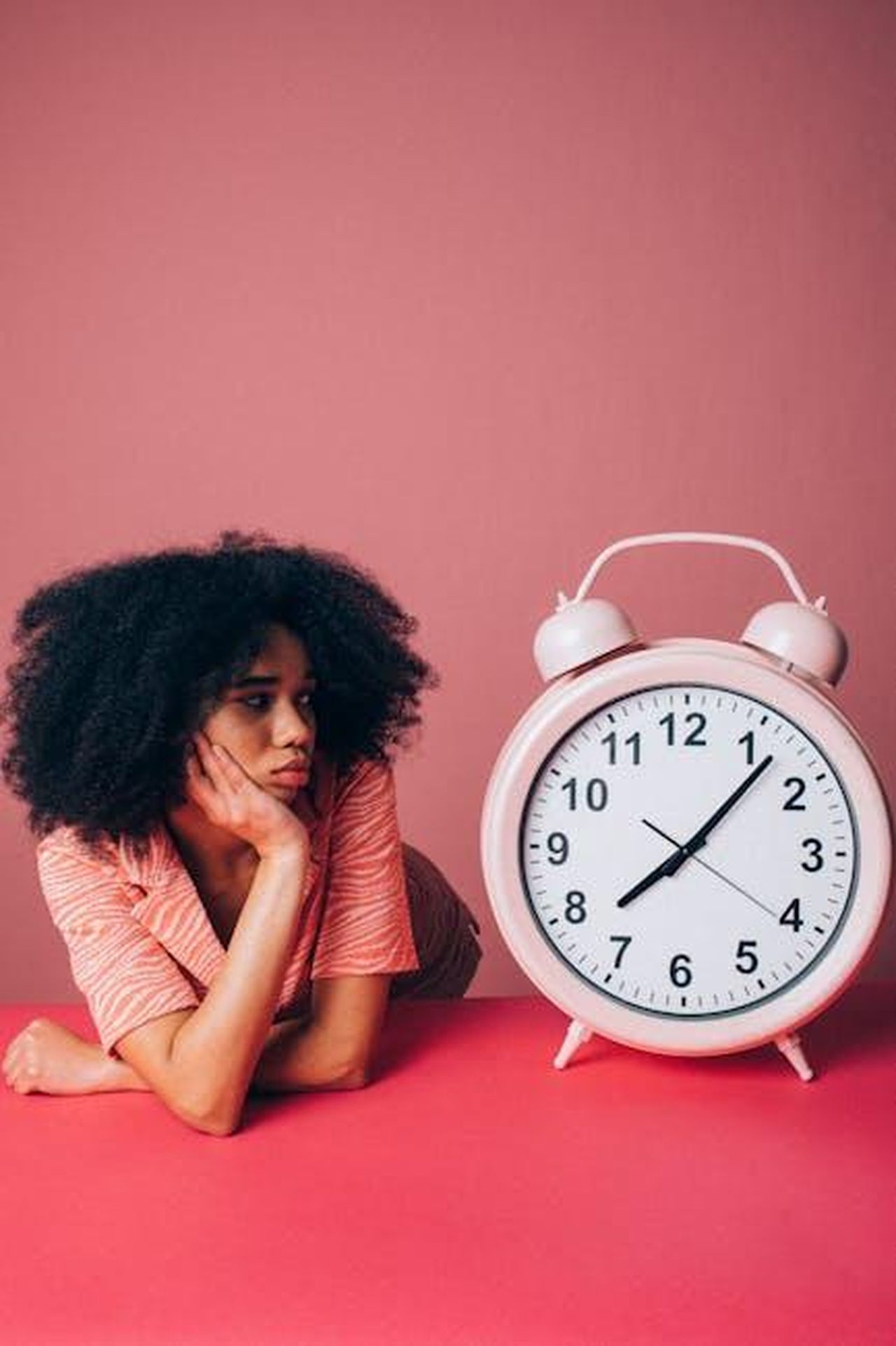 Limpatienza_di_Meghan_eZ3Q7EuiaB - Confidenze24 Source: KoolShooters - pexels - Woman in a pink shirt leans on table with a large alarm clock, conveying impatience.