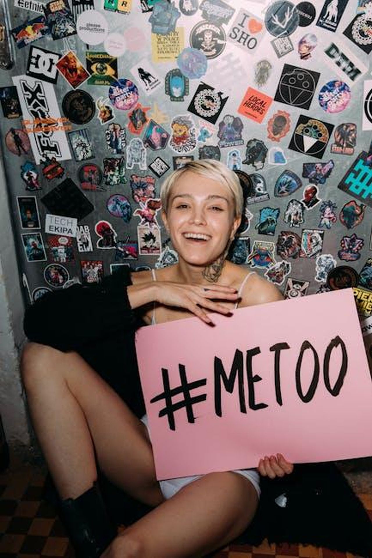 Limpatto_del_movimento_MeToo_GuQEzyGWFl - Confidenze24 Source: Viktoria Slowikowska - pexels - A woman smiling while holding a #MeToo placard in front of a wall covered with stickers.