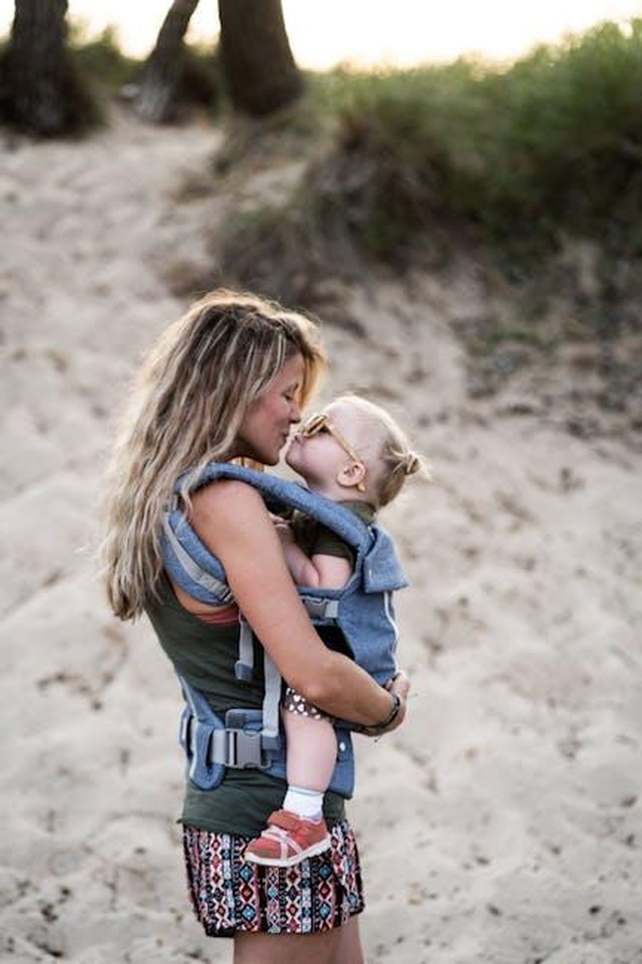 Limpatto_della_maternit_su_Keke_Palmer_brjW38zsQH - Confidenze24 Source: Josh Willink - pexels - A loving mother kisses her toddler in a baby carrier on a sandy beach, capturing a tender parenting moment.
