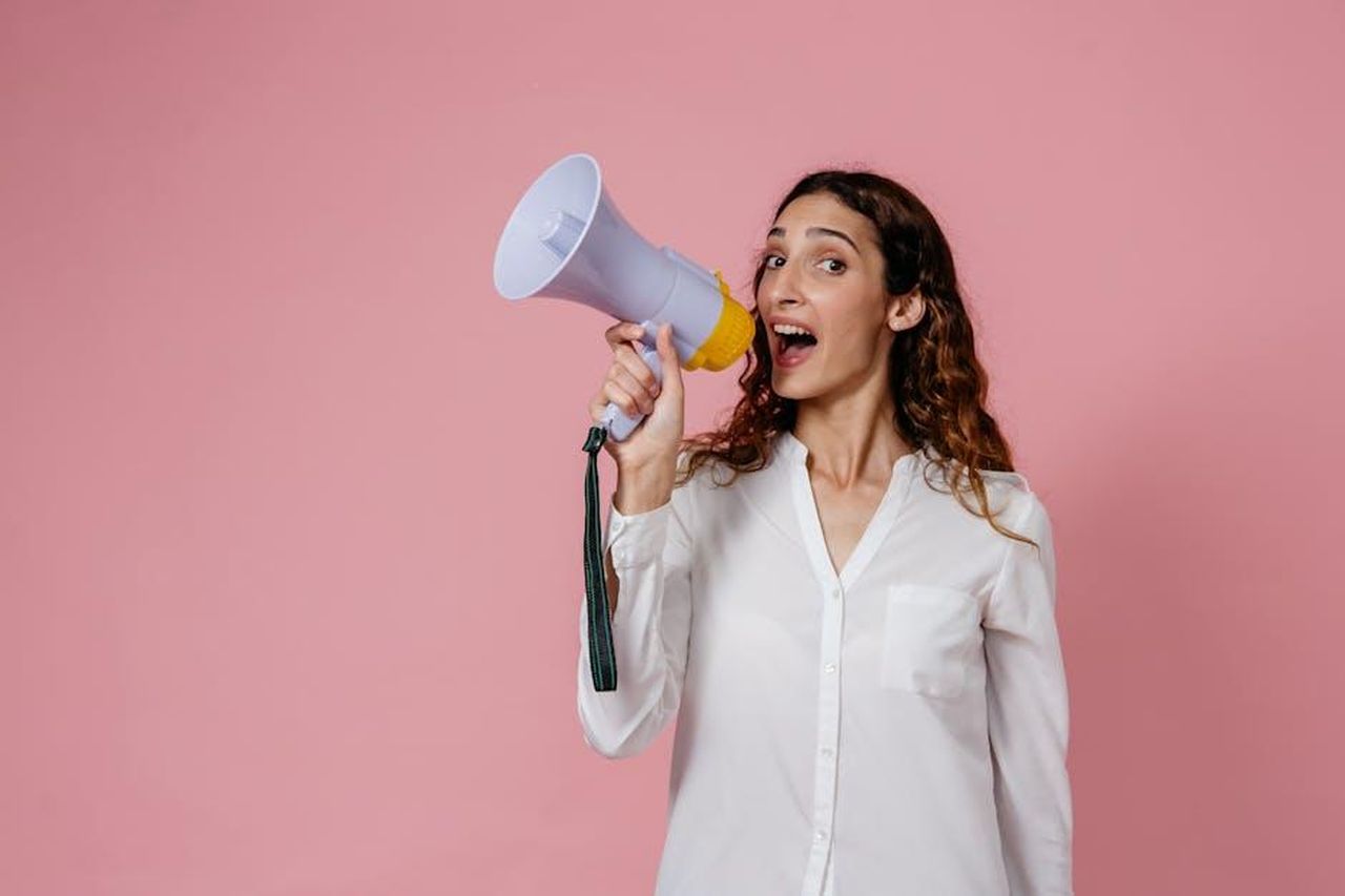 Source: Pavel Danilyuk - pexels - Woman in white shirt holding megaphone against a pink background, confidently speaking.