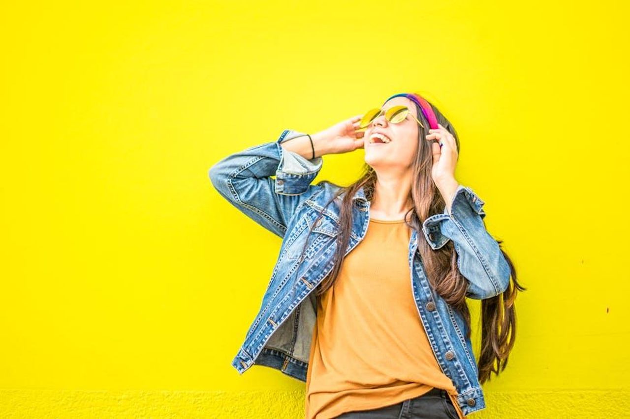 Lo_stile_di_Kate_al_match_a6p65Vohmo - Confidenze24 Source: juan mendez - pexels - Smiling woman in sunglasses stands against vibrant yellow wall, radiating happiness.