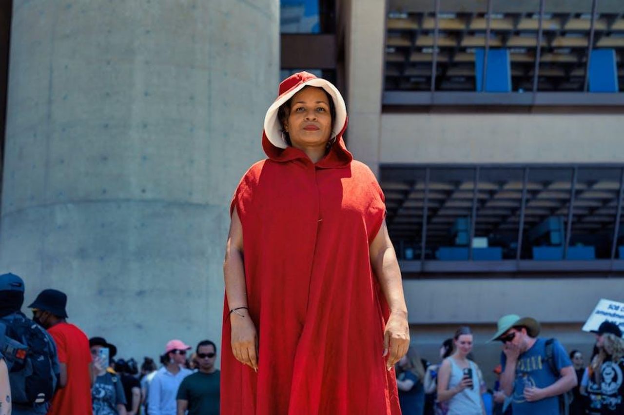 Lultima_stagione_di_The_Handmaids_Tale_e_il_legame_con_la_figlia_uHBqYG8WnG - Confidenze24 Source: Alfo Medeiros - pexels - A person in a handmaid costume stands confidently outside a modern building during a cosplay event.