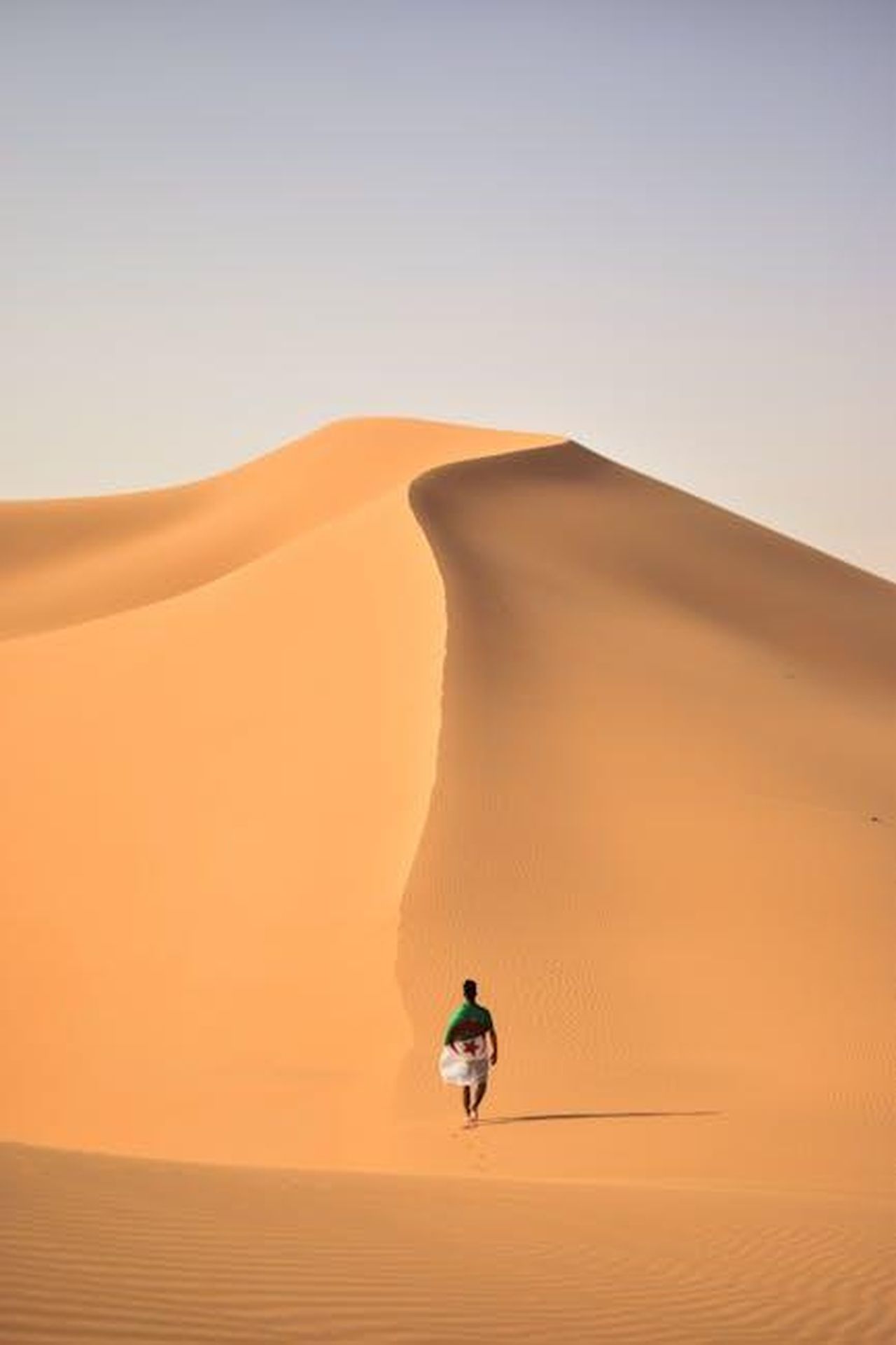 Maura_lascia_il_Regno_Unito_amid_scandal_ZOyg6lr86U - Confidenze24 Source: Amine M'siouri - pexels - A lone traveler walking over the vast, golden sand dunes of the Sahara Desert in Algeria.