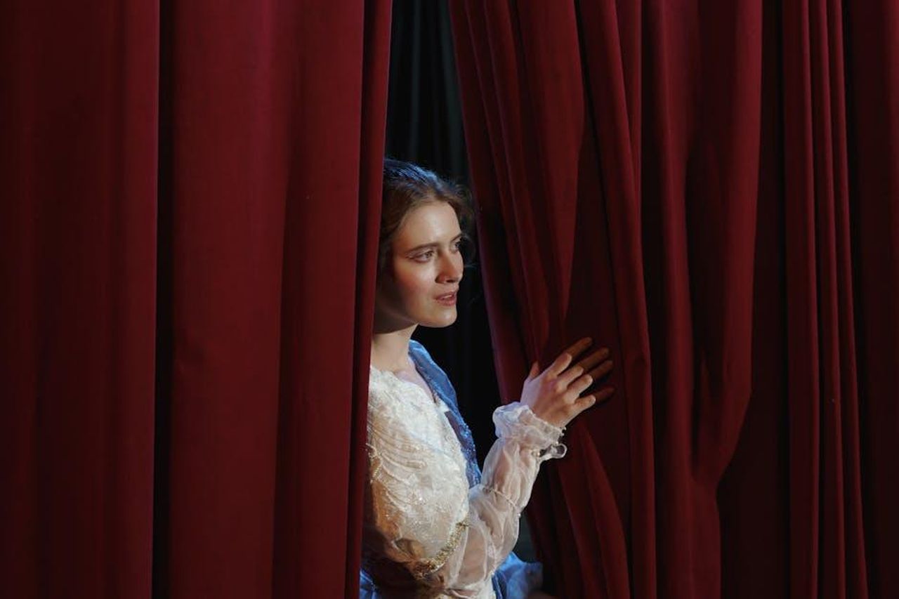 Maura_si_innamora_di_Danny_23HdpGKz1k - Confidenze24 Source: cottonbro studio - pexels - A woman in costume peeks through red theater curtains in a dramatic scene.