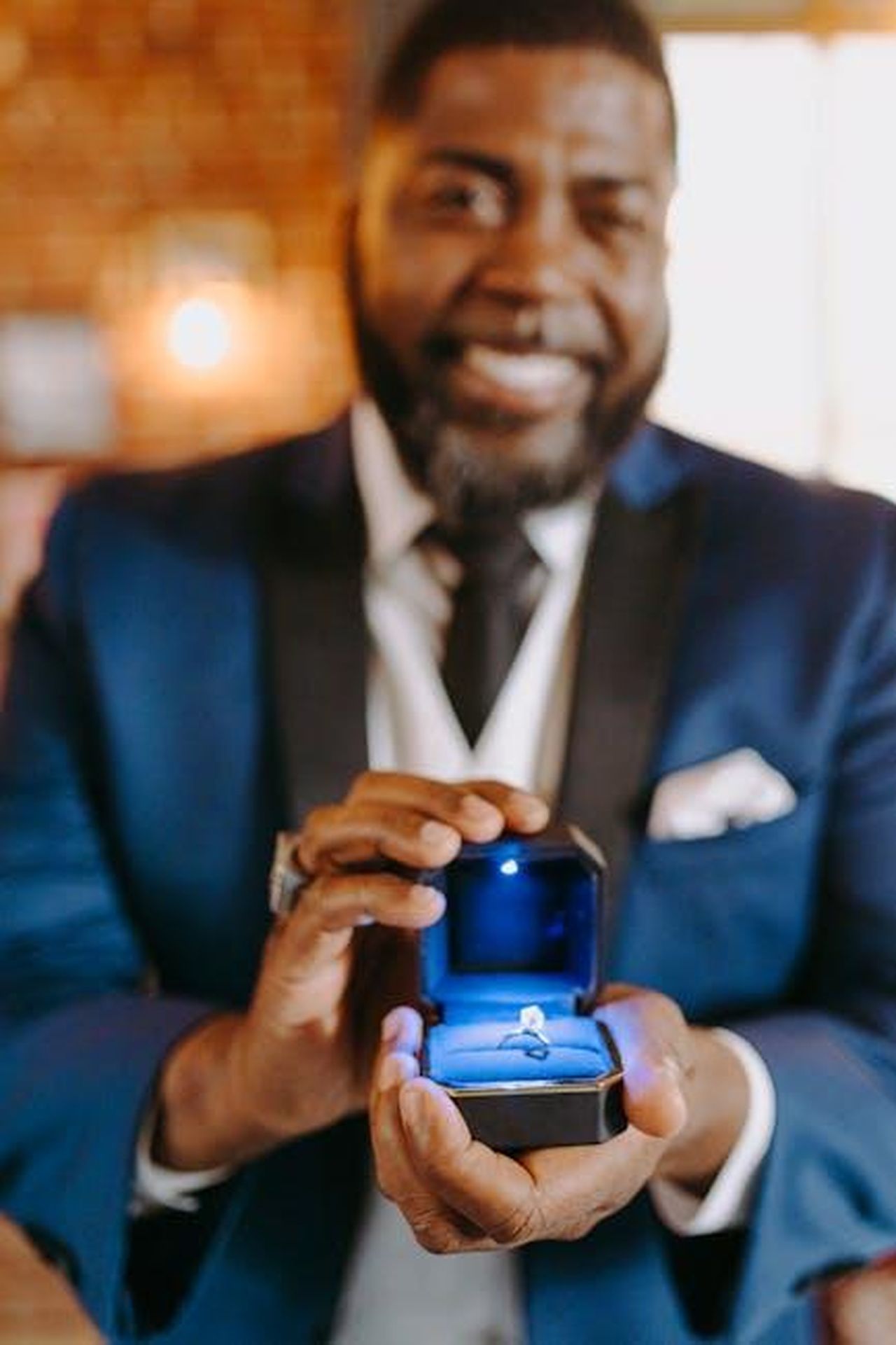 Miller_e_Garner_pianificano_di_sposarsi_4Bi2kZWBHf - Confidenze24 Source: RDNE Stock project - pexels - Man proposing with a ring in a blue box, wearing a suit, capturing a romantic moment.