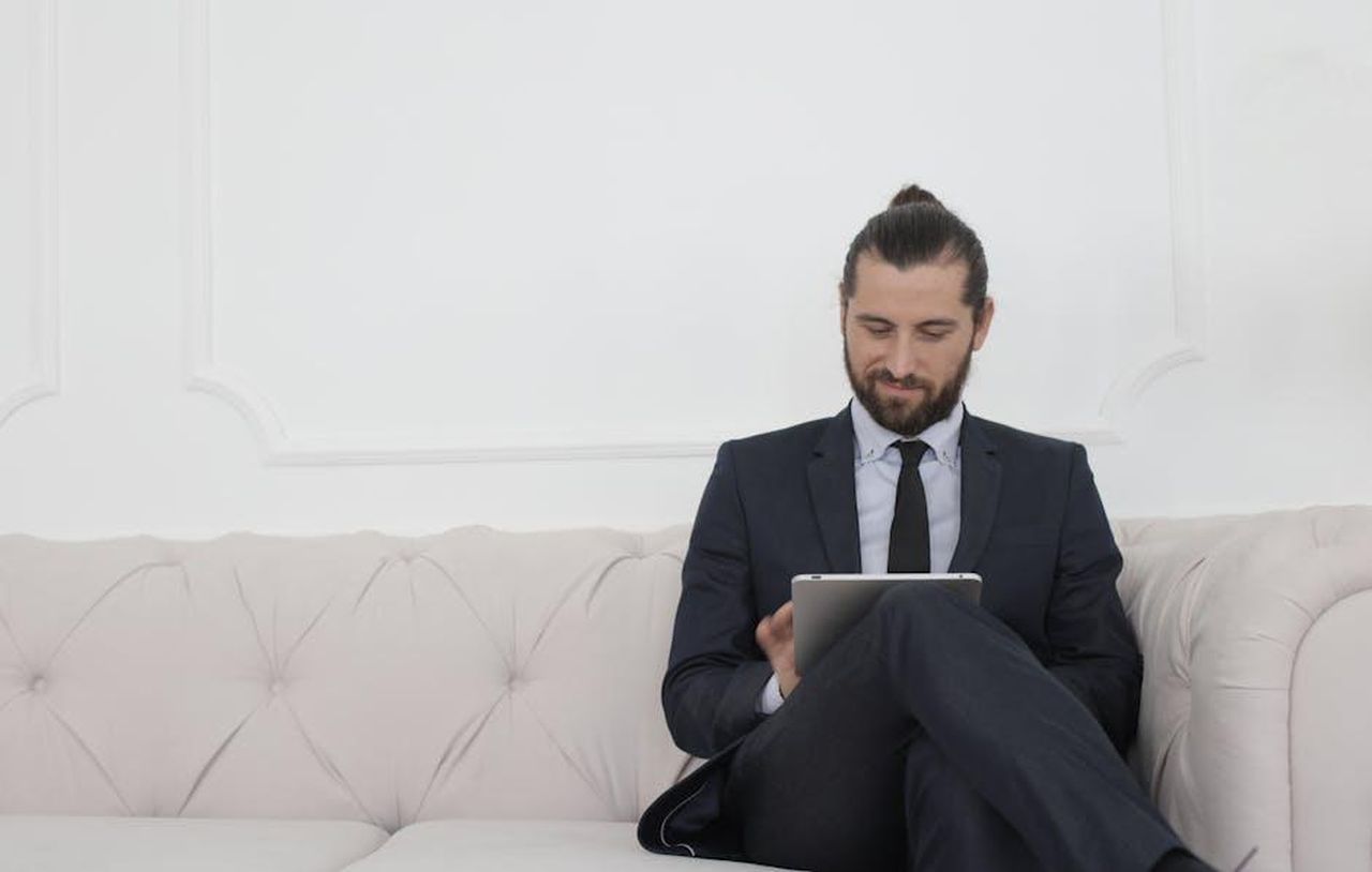 Non_si_identifica_come_influencer_o_tradwife_VfN9StPCzn - Confidenze24 Source: Andrea Piacquadio - pexels - A businessman in a suit sitting on a sofa using a tablet, smiling.