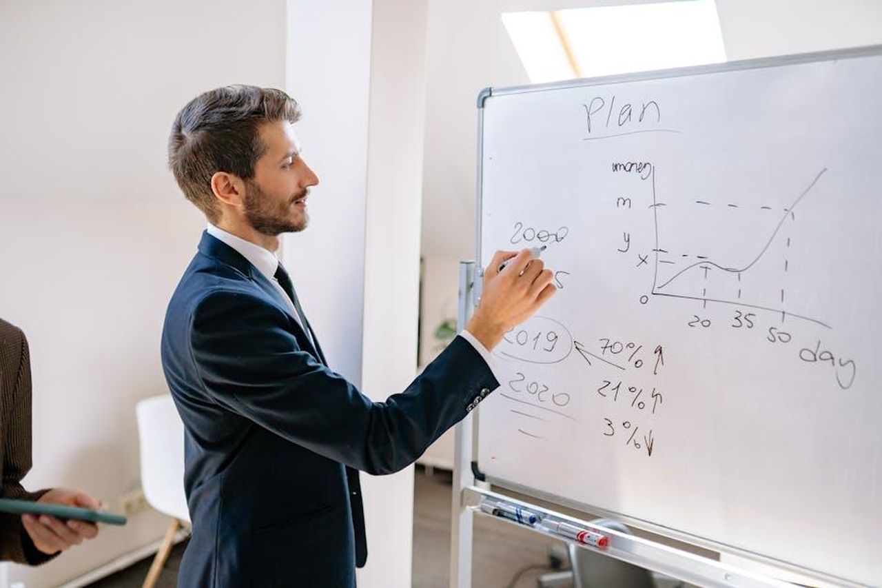 Piani_Futuri_di_Hoda_Kotb_6001flXHhT - Confidenze24 Source: Yan Krukau - pexels - A businessman in a suit writes financial data on a whiteboard during an office planning session.