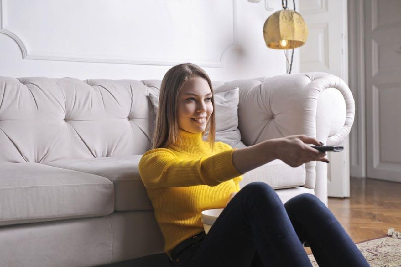 Possibile_Spinoff_di_The_Neighborhood_rhbjIB7BJW - Confidenze24 Source: Andrea Piacquadio - pexels - Joyful millennial female in casual clothes with bowl of snack using remote controller while sitting on floor leaning on sofa and watching movie in cozy light living room with luxury interior