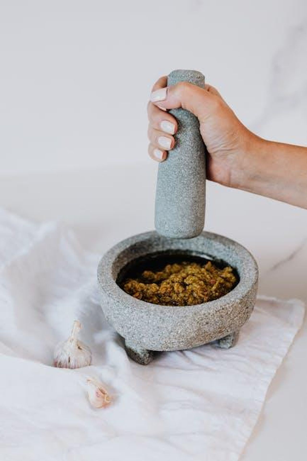 Preparazione_del_pesto_per_il_sandwich_di_salumi_WmXrHCCA0Q - Confidenze24 Source: Photo By: Kaboompics.com - pexels - A hand using a stone mortar and pestle to grind ingredients with garlic cloves nearby.