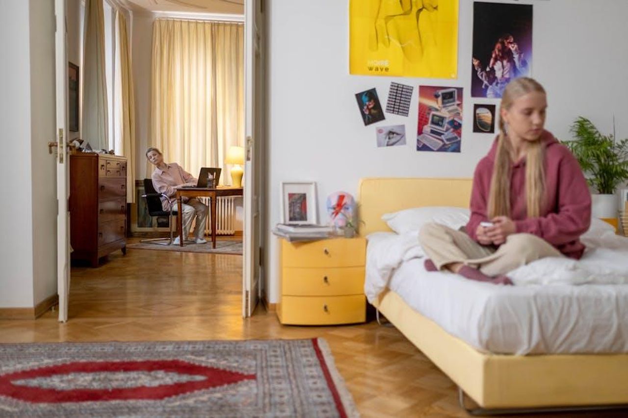Quali_sono_gli_effetti_a_lungo_termine_della_registrazione_dei_teenager_j9JH3NFMH7 - Confidenze24 Source: cottonbro studio - pexels - Teenager sitting on bed, with parent working in next room. Cozy home interior.