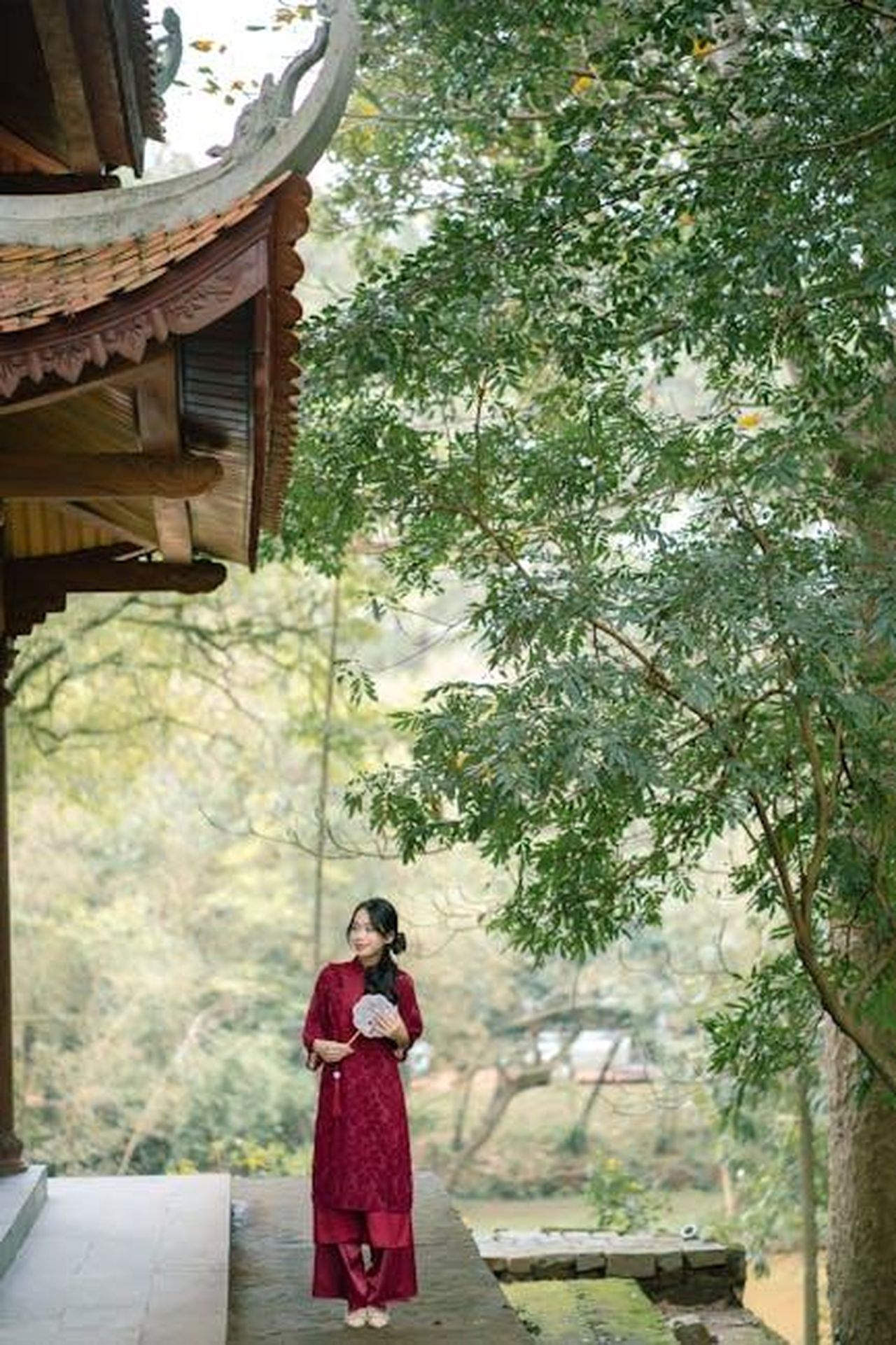 Reazioni_dei_fan_e_commenti_divertenti_KNrJ3GtORK - Confidenze24 Source: Anh Quốc - pexels - A woman in red traditional clothing holds a fan outside an Asian temple surrounded by greenery.