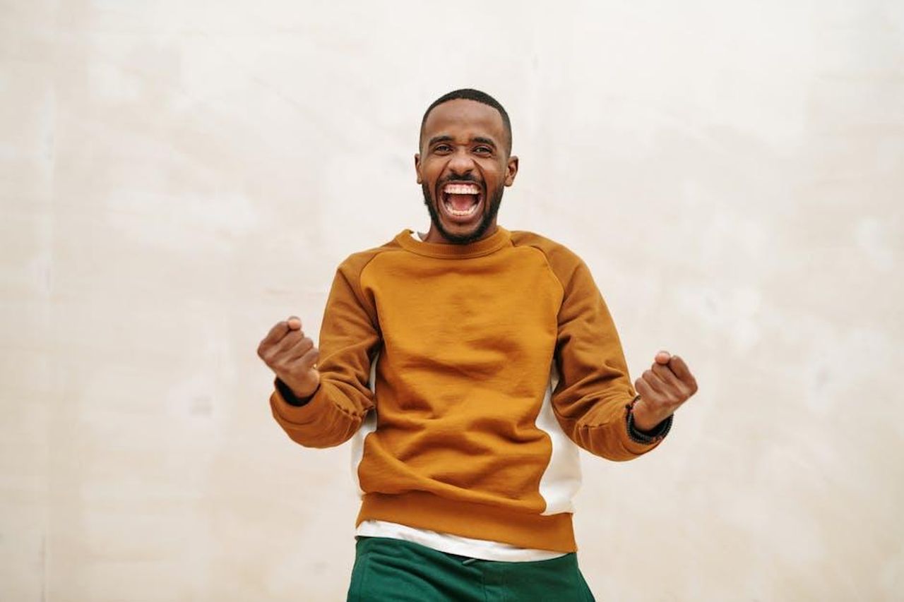 Reazioni_di_Jennifer_Lopez_al_gossip_ULnfkaNfV6 - Confidenze24 Source: Diva Plavalaguna - pexels - A joyful man with a beard in a sweater, expressing excitement and celebration indoors.