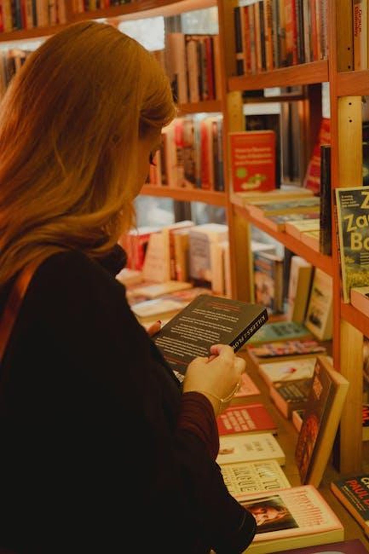 Reazioni_e_commenti_sul_libro_WWqYTMCQ56 - Confidenze24 Source: Eyüpcan Timur - pexels - Woman browsing a book in a cozy bookstore filled with shelves of books.
