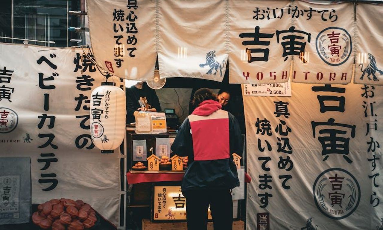 Recensioni_e_testimonianze_sul_CeraVe_Skin_Renewing_Night_Cream_wJ1hliqUB0 - Confidenze24 Source: gabriele donofrio - pexels - A traditional Japanese street food stall with banners and a customer buying snacks.