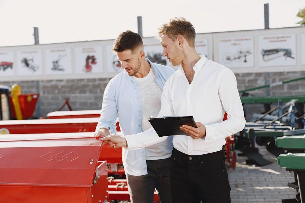 Recensioni_entusiastiche_dei_clienti_xk3SMWEqJa - Confidenze24 Source: Gustavo Fring - pexels - Two men discussing agricultural equipment purchase at a dealership, one holding a clipboard outside.