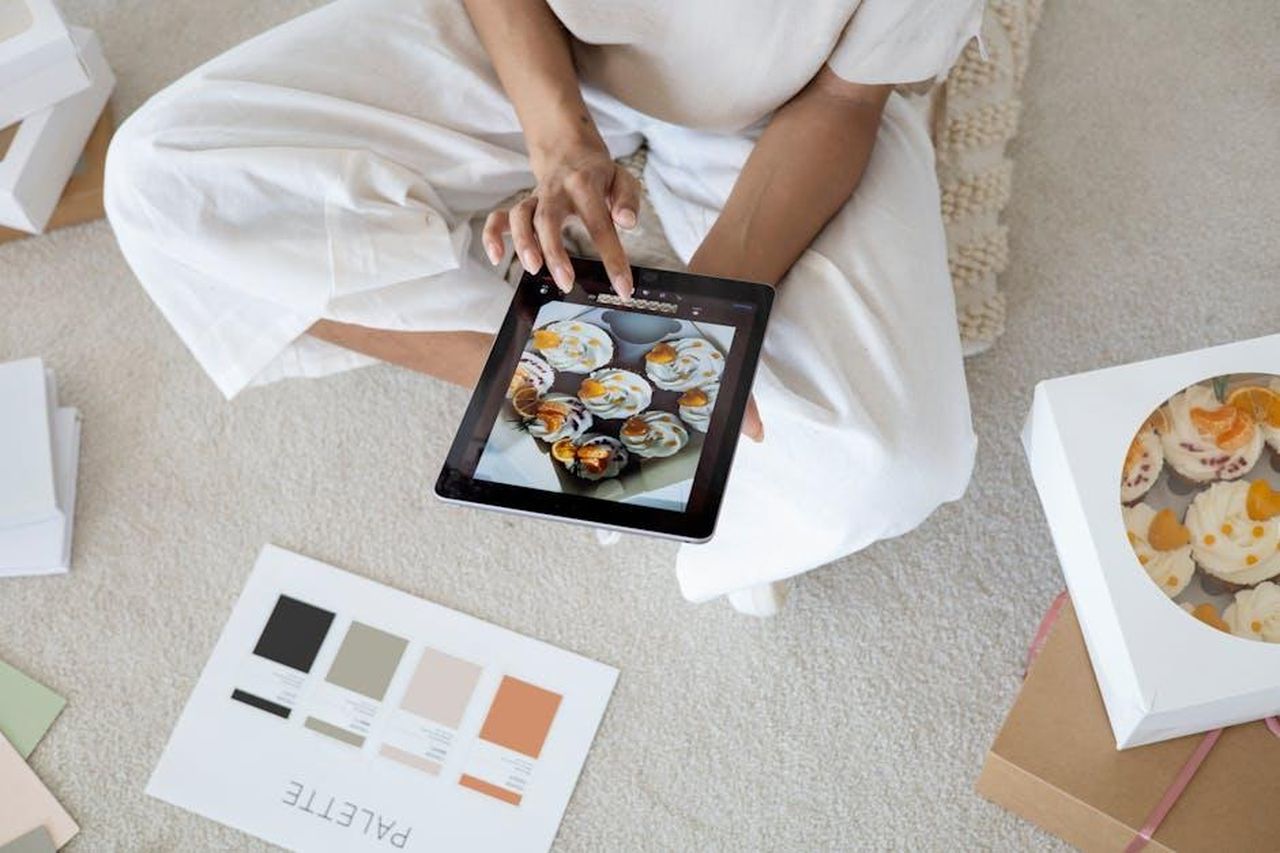 Recensioni_entusiastiche_su_Instagram_Q9uwWeKLop - Confidenze24 Source: Anna Nekrashevich - pexels - An individual selects cupcakes via tablet, surrounded by palette and cupcake box indoors.