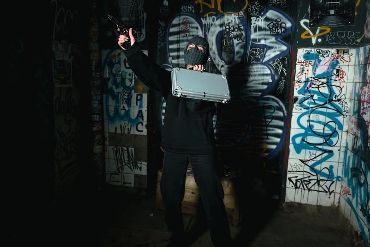Richiesta_di_Scuse_e_Minacce_di_Esporre_la_Verit_YSEi0keArr - Confidenze24 Source: MART PRODUCTION - pexels - A masked person holding a briefcase and gun in a graffiti-covered alley at night.