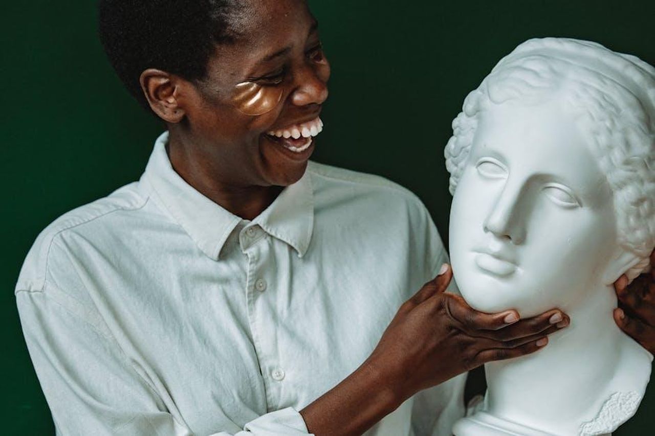 Ridi_per_Allontanare_lo_Stress_Ma7NNJGom0 - Confidenze24 Source: ROMAN ODINTSOV - pexels - Happy woman in skincare regimen smiling at sculpture indoors.